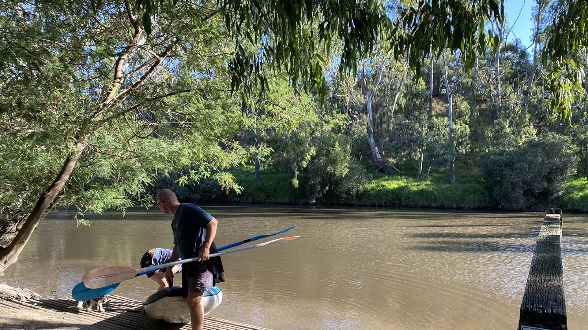 A person in a dark t-shirt and cap lifts the front of a kayak out of a river. They hold a paddle in their left hand while another person bends down to lift up the back end of the kayaki