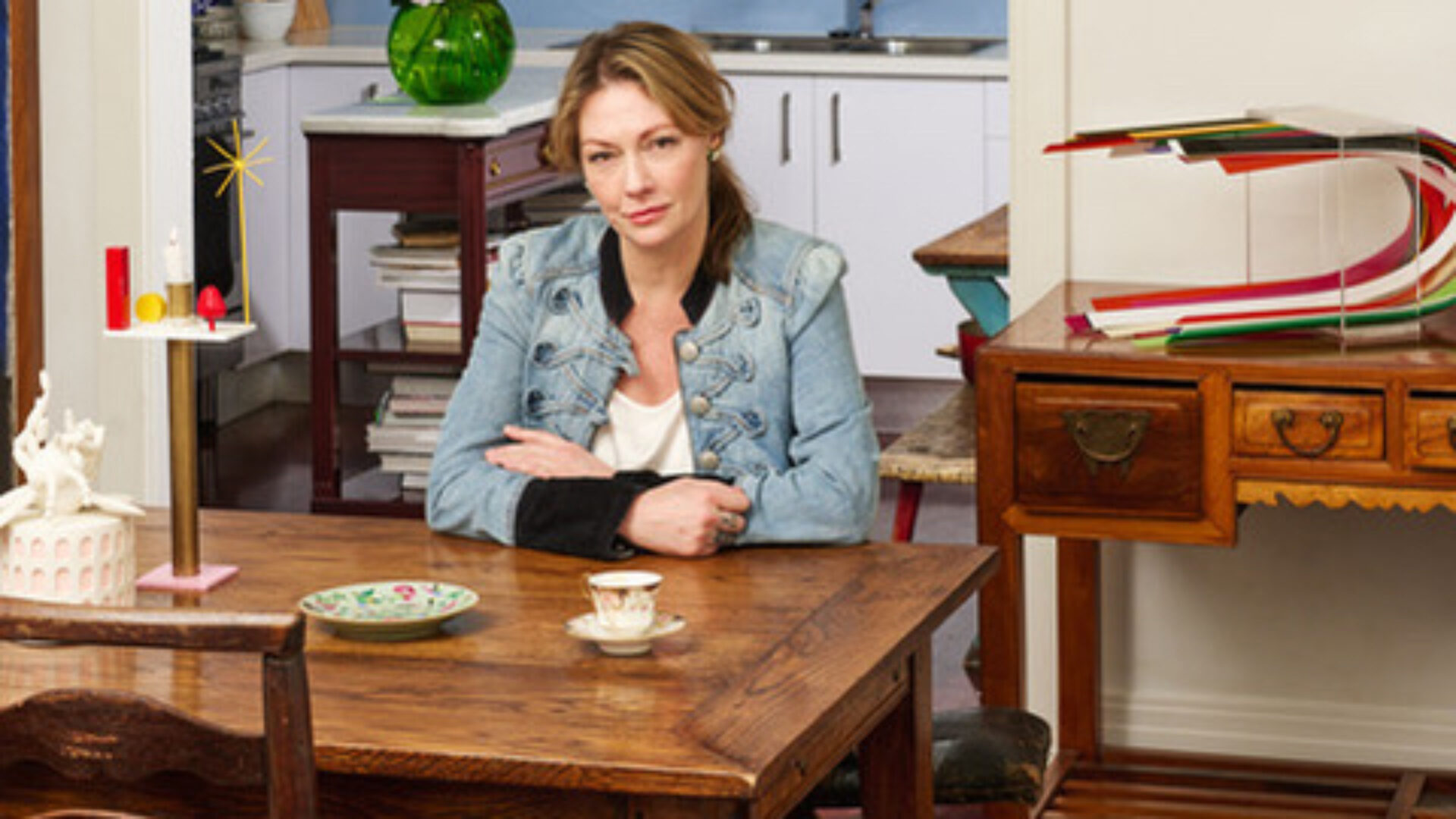 Gallerist and curator Sarah Fletcher wearing a denim jacket seated at a table surrounded by works of artists she represents