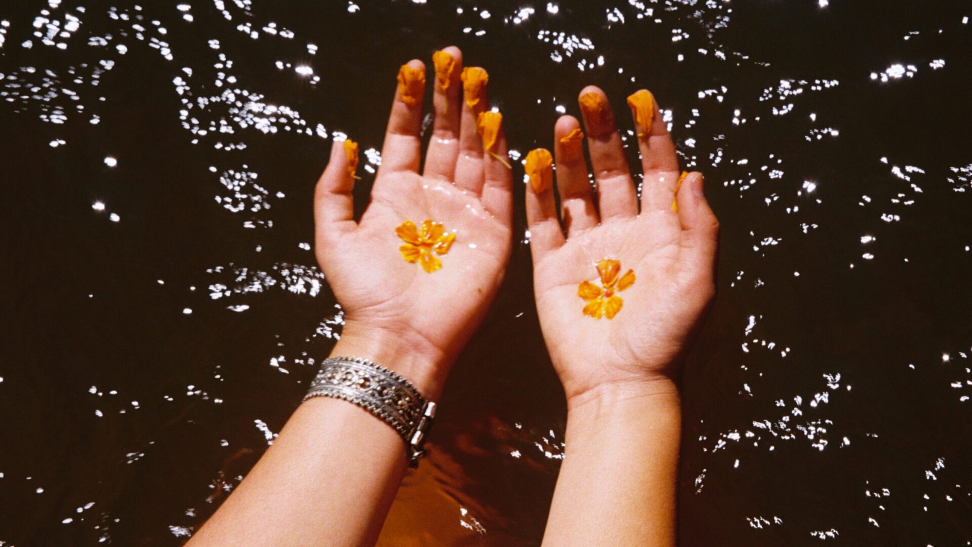 The image shows two hands held with their palms up above a body of water. In each palm is a small yellow flower and each finger is decorated with yellow flower petals. The left arm wears a silver watch and has a small rose tattoo visible.