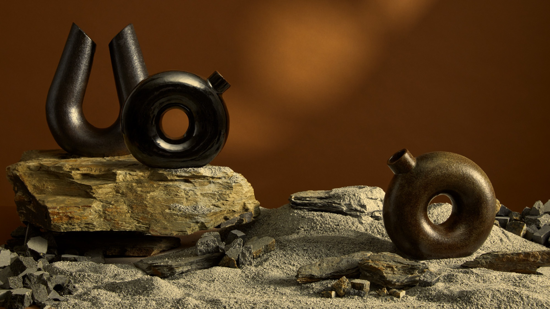 Two ceramic forms and three earth toned glazes of sculptural decanters on granite sand and a large piece of slate, surrounded by bluestone gravel against a brown background with dappled light