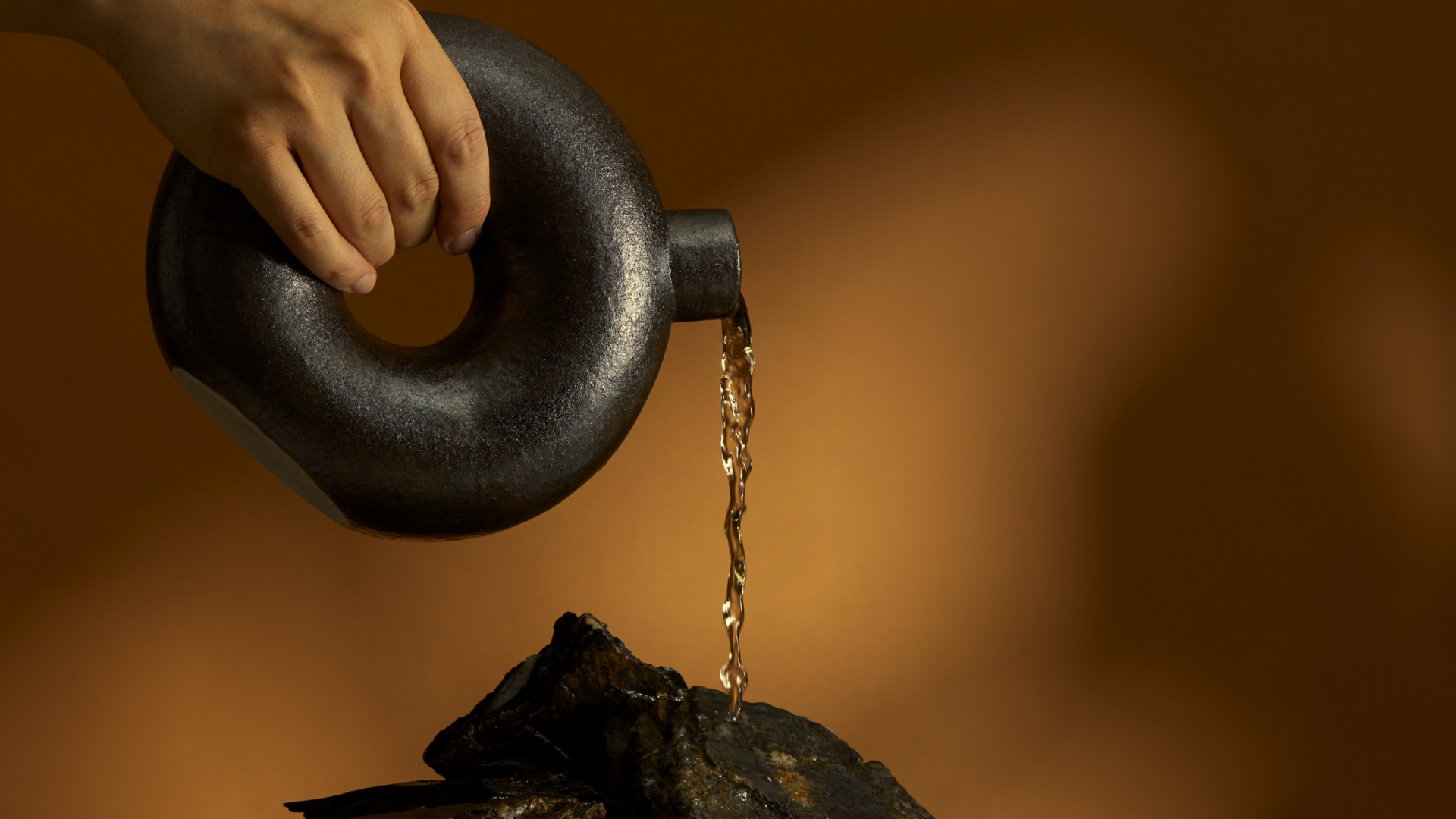 Photo of a ceramic ring shaped vessel with a spout pouring white wine onto slate against a brown backdrop with dappled light