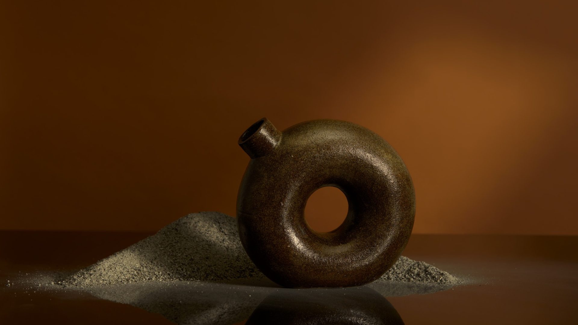 Ring shaped ceramic decanter with a brown textured glaze in front of a pile of granite sand