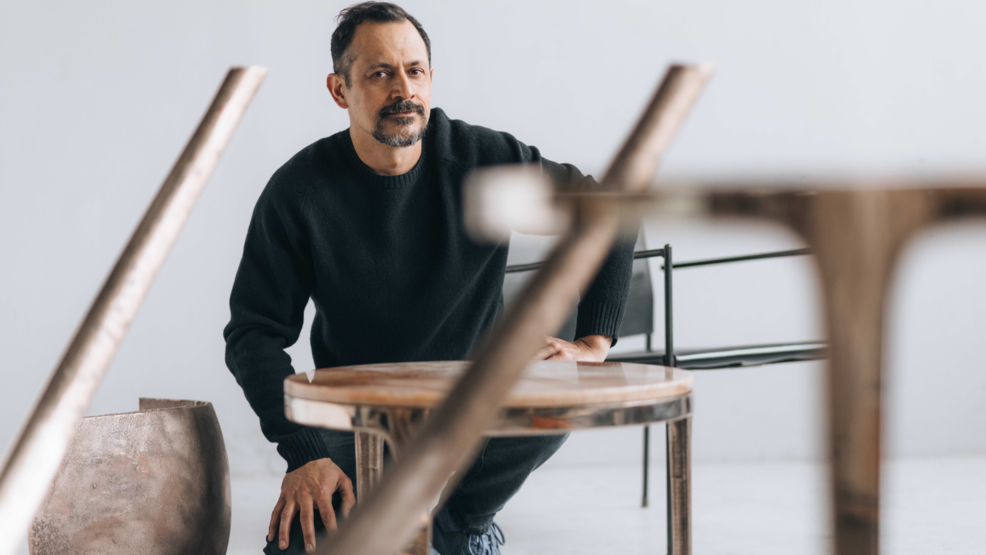 Designer Daniel Barbera kneeling in his studio surrounded by his own works including a bronze and marble s ide table and a leather dining chair