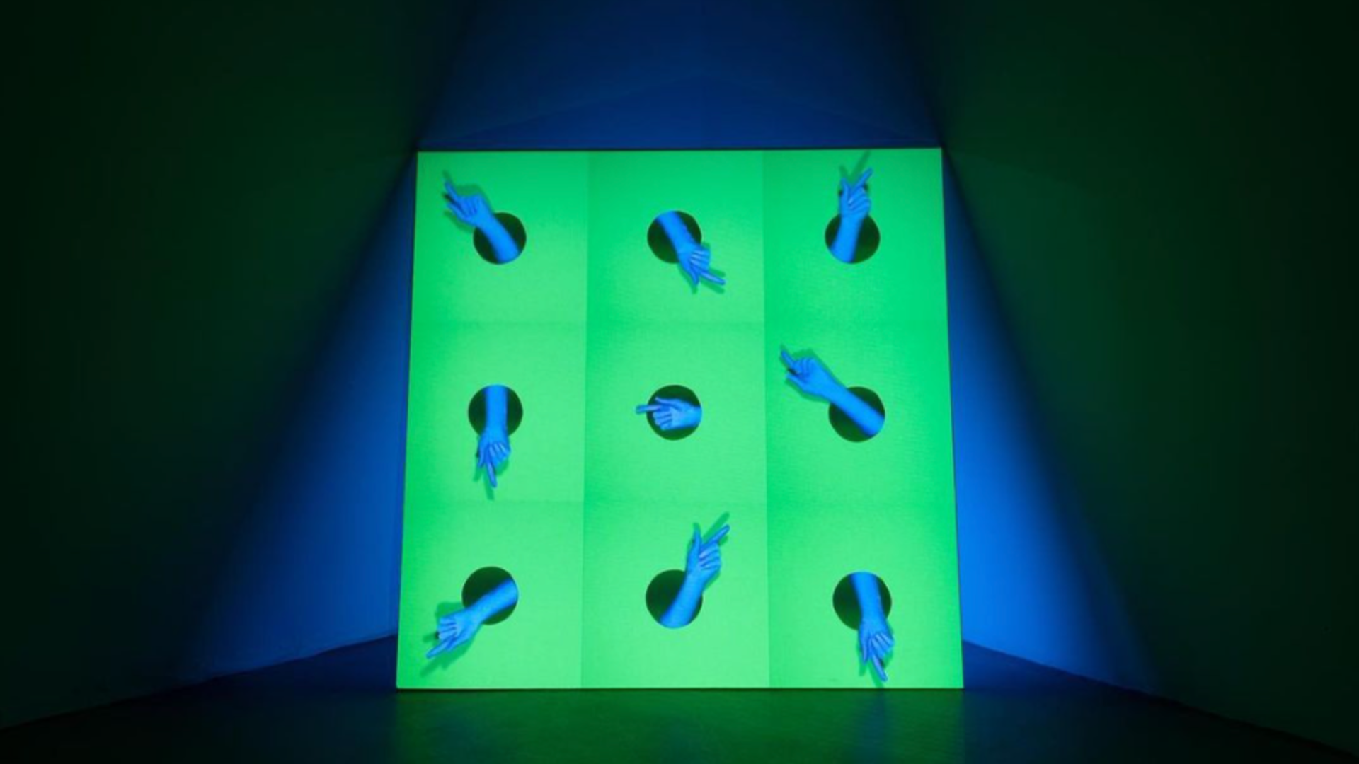 Photo of a digital video collage installation showing a green background and series of blue gloved hands projected onto a screen.